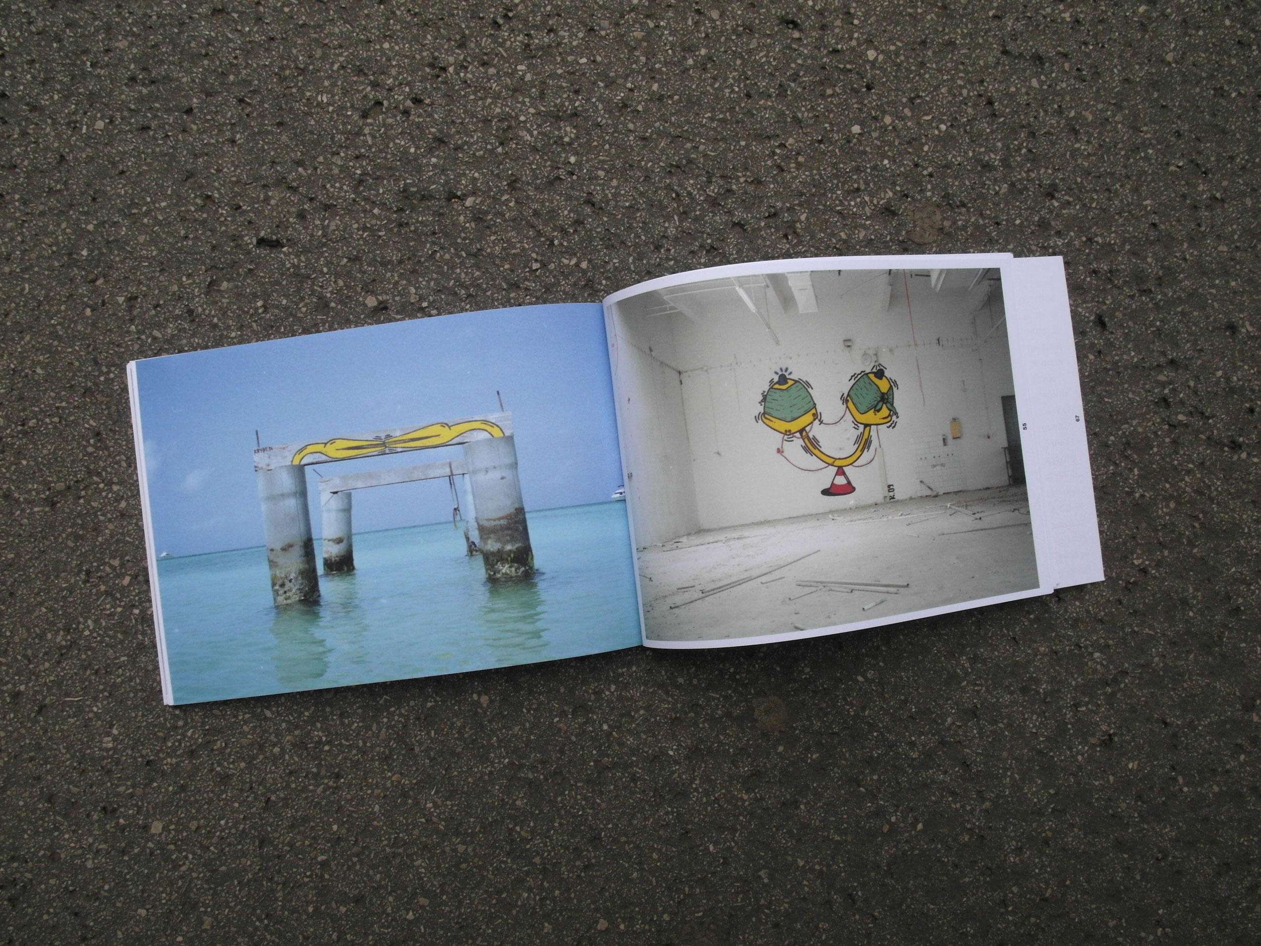 Open book lying on the street. Left double page: Photo of old jetty construction on the seashore. Partly painted with yellow/black motif. Right double page: Photo inside abandoned warehouse. Huge motif painted on wall in yellow/black.